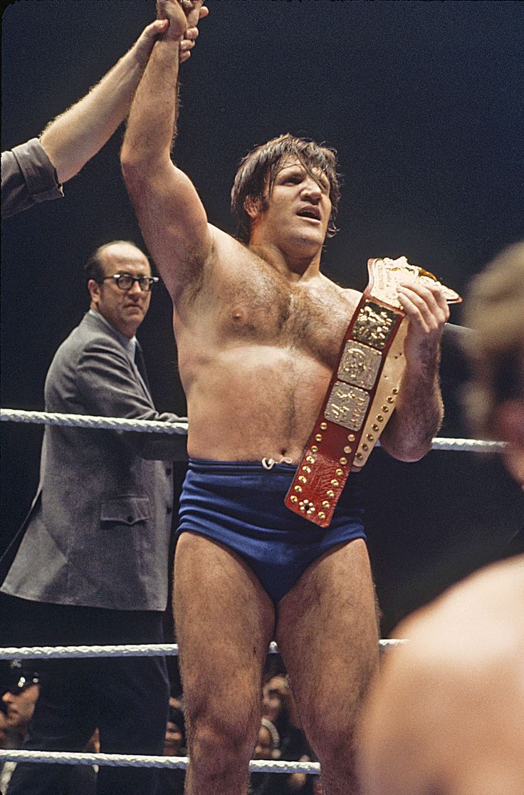 a man in blue wrestling trunks holding up a belt with one hand and two other hands