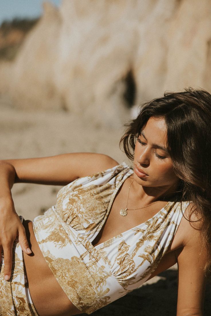 a woman in a white and gold swimsuit laying on the beach with her hand on her hip