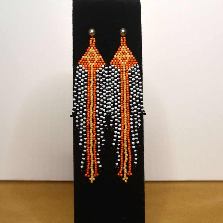 Handmade, one of a kind earrings are made with glass seed beads in gold, orange, black and white. They are fitted with a post setting. The earrings are 4 1/2 inches in length. Orange Beaded Earrings With Black Beads, Orange Dangle Earrings With Black Beads, Orange Dangle Jewelry With Black Beads, Orange Dangle Beaded Earrings With Black Beads, Orange And Black Beads Dangle Earrings, Orange Earrings With Black Beads As Gift, Orange Earrings With Black Beads For Gift, Orange Beaded Drop Earrings For Pierced Ears, Unique Orange Earrings With Dangling Beads