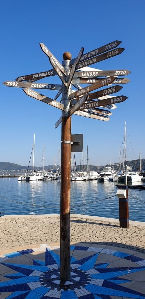 a wooden pole with many signs on it near the water and boats in the background