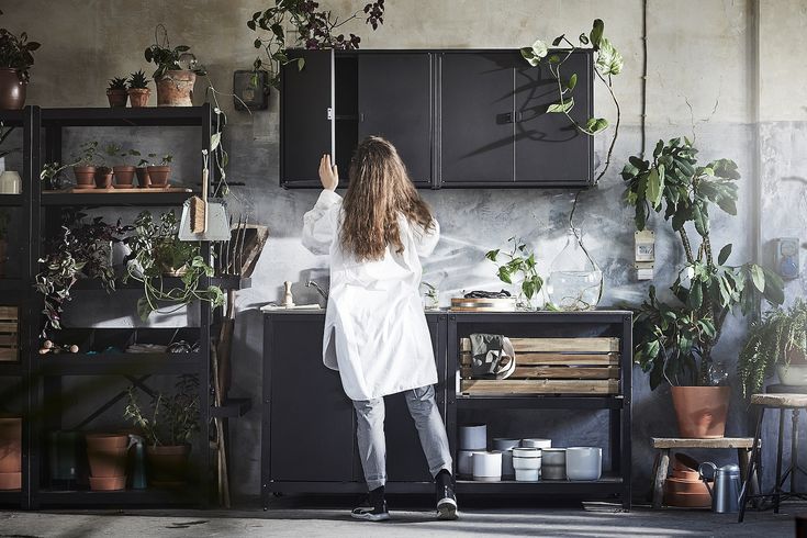 a woman standing in front of a kitchen counter with potted plants on the shelves