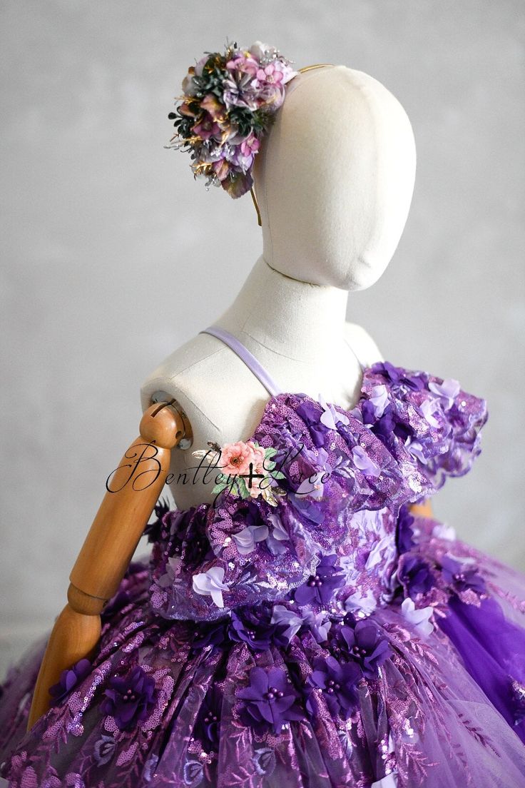 Our beautiful "Amethyst" gown is made with a beautiful 3D floral fabric. Purple perfection. We've added a beautiful on bodice flutter detail which truly adds to her sweet charm. We have added a very flexible back which features shoulder ties which add to the ease of size adjustment and a full elastic back- which lends to her ease of size adjustment as well. Typically with our elastic back you can add an additional 1-3 inches of room into this dress. Her skirting features yards of bridal soft tul Purple Princess Dress With Floral Applique For Parties, Lavender Fitted Princess Dress For Dress-up, Fitted Fairy Dress With Ruffles For Wedding, Purple Fitted Ball Gown Princess Dress, Fitted Tulle Fairy Dress For Garden Party, Fitted Princess Dress With Ruffles For Garden Party, Pageant Gown With Ruffles And Fitted Bodice, Purple Fitted Tulle Ball Gown, Elegant Purple Dress-up Gown