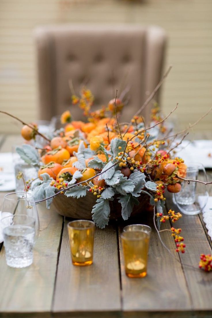 an arrangement of oranges and greenery is displayed on a wooden table with glasses