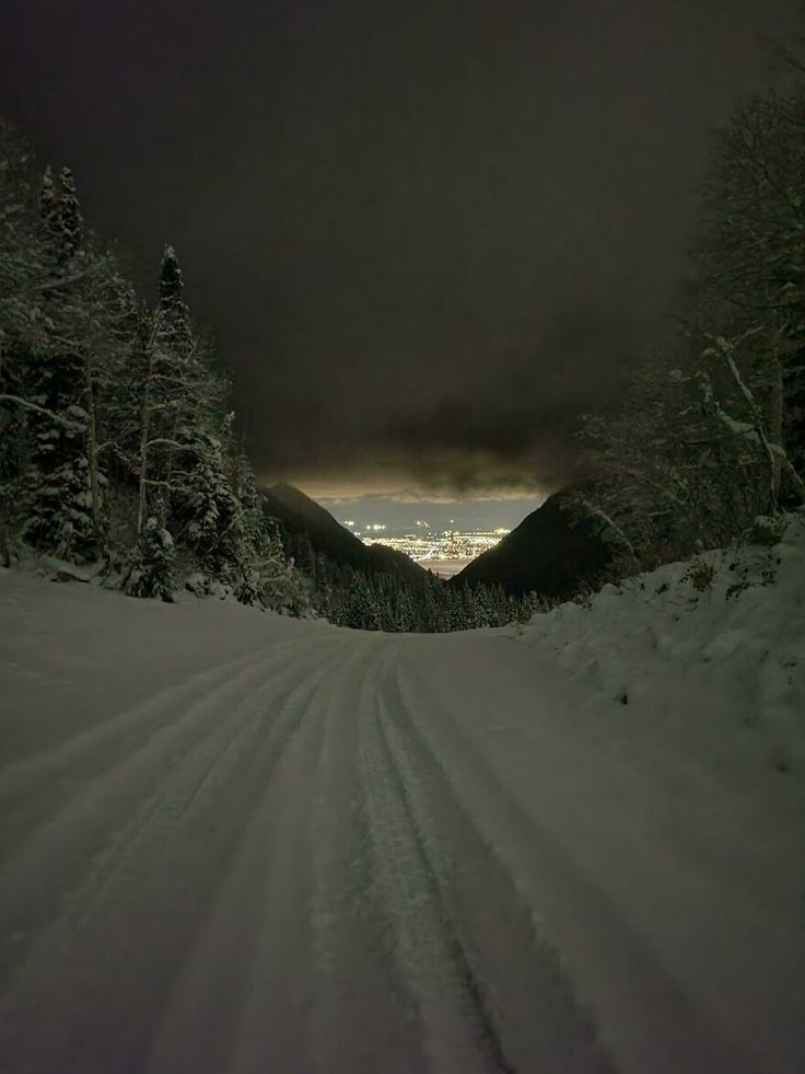 a snow covered road with trees on both sides and dark clouds in the sky above