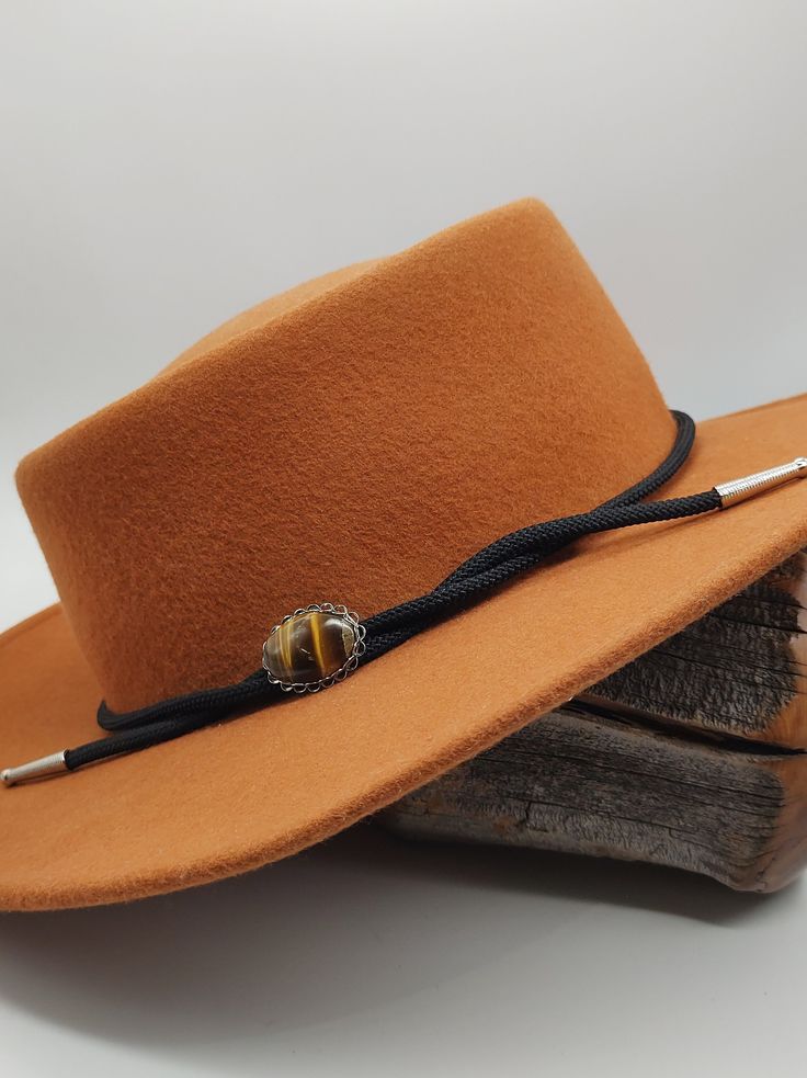 Elevate your cowboy hat style with our exquisite handcrafted hat band featuring a captivating oval Tigers Eye stone as its centerpiece. Each stone is meticulously polished to perfection, showcasing its natural beauty and warm glow. Designed to add a touch of elegance to your western look, this hat band is adorned with an oval silver-tone setting, creating a striking contrast against the rich hue of the stone. The band is crafted from black vegan cording, accented with fancy silver-tone "stacked" end caps. Hat bands are more than just accessories; they're a form of personal expression, reflecting your unique style and personality. As someone who grew up on the ranch, I understand the significance of these adornments. Adjustable to fit different hat sizes, this hat band offers versatility an Adjustable Country Style Top Hat For Outdoor, Adjustable Flat Brim Country Fedora, Country Style Fedora With Adjustable Curved Brim, Adjustable Wide Brim Country Style Top Hat, Country Style Adjustable Fedora With Curved Brim, Adjustable Wide Brim Top Hat For Country Events, Country Style Adjustable Fedora With Flat Brim, Country Style Adjustable Wide Brim Top Hat, Western Hat Bands For Country Events