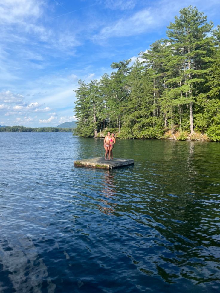 a man standing on a dock in the middle of a body of water with trees behind him