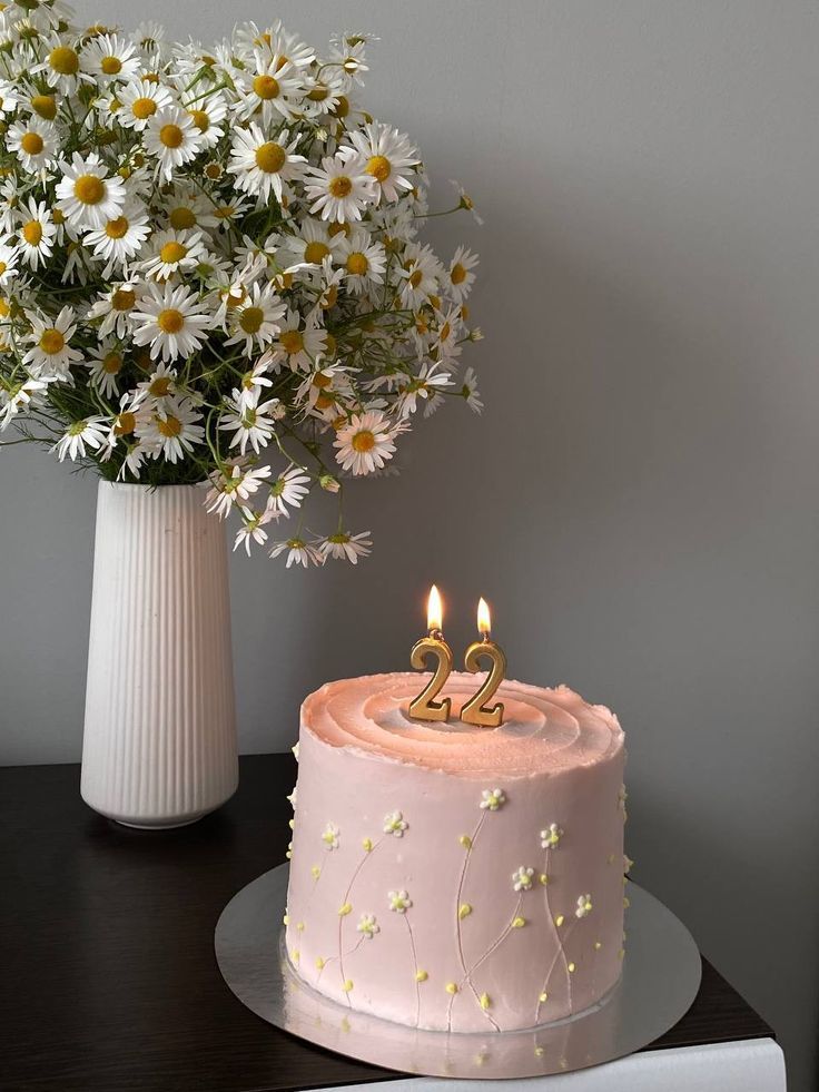 a pink birthday cake with two candles on it and daisies in a vase next to it