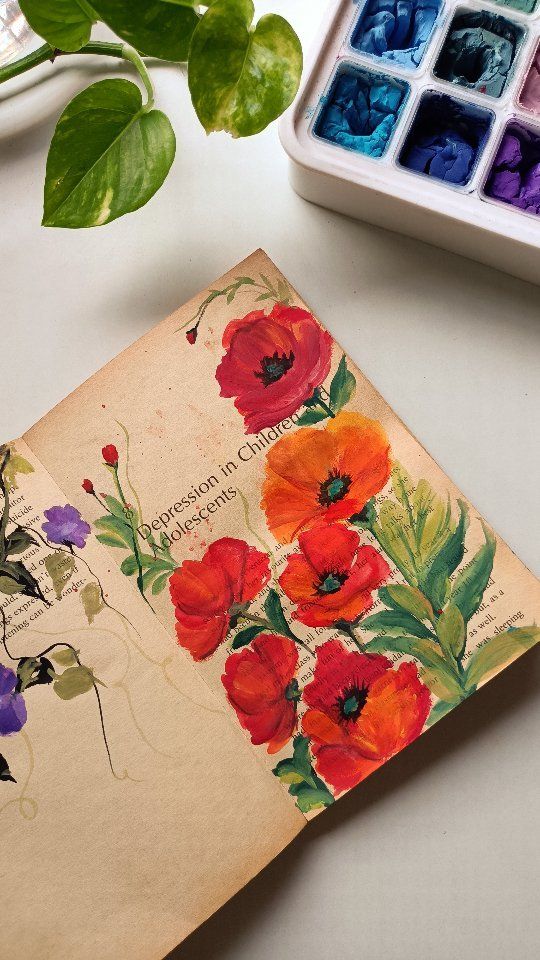 an old book with flowers painted on it next to some paintbrushes and a potted plant