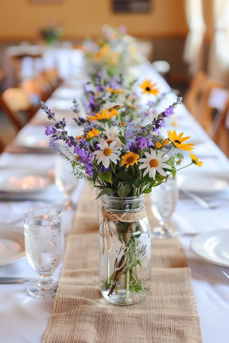 a long table with flowers in a mason jar on the centerpiece and place settings