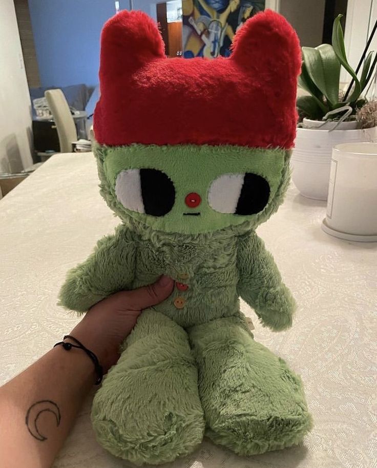 a green stuffed animal sitting on top of a table next to a person's hand