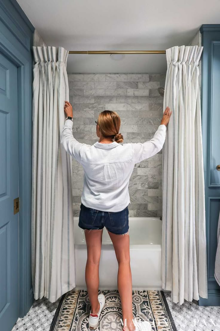 a woman standing in front of a bathtub with curtains on the window sill