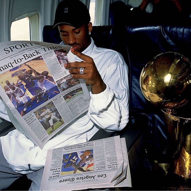 a man sitting in a chair reading a newspaper while holding a gold trophy and looking at his cell phone