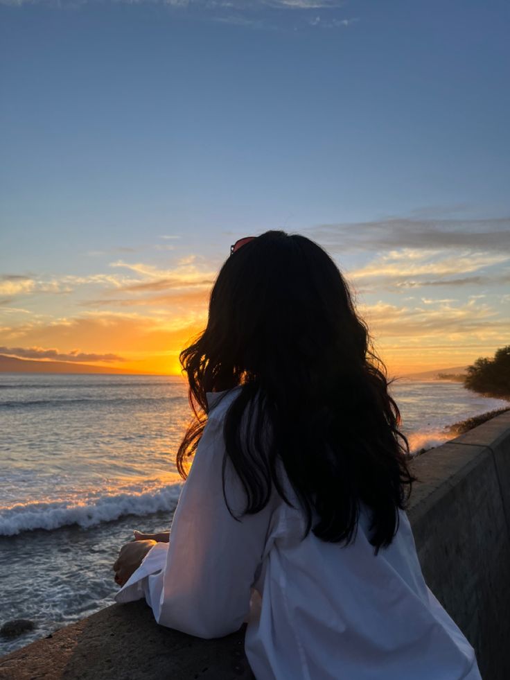 a woman sitting on the edge of a wall looking out at the ocean while the sun sets