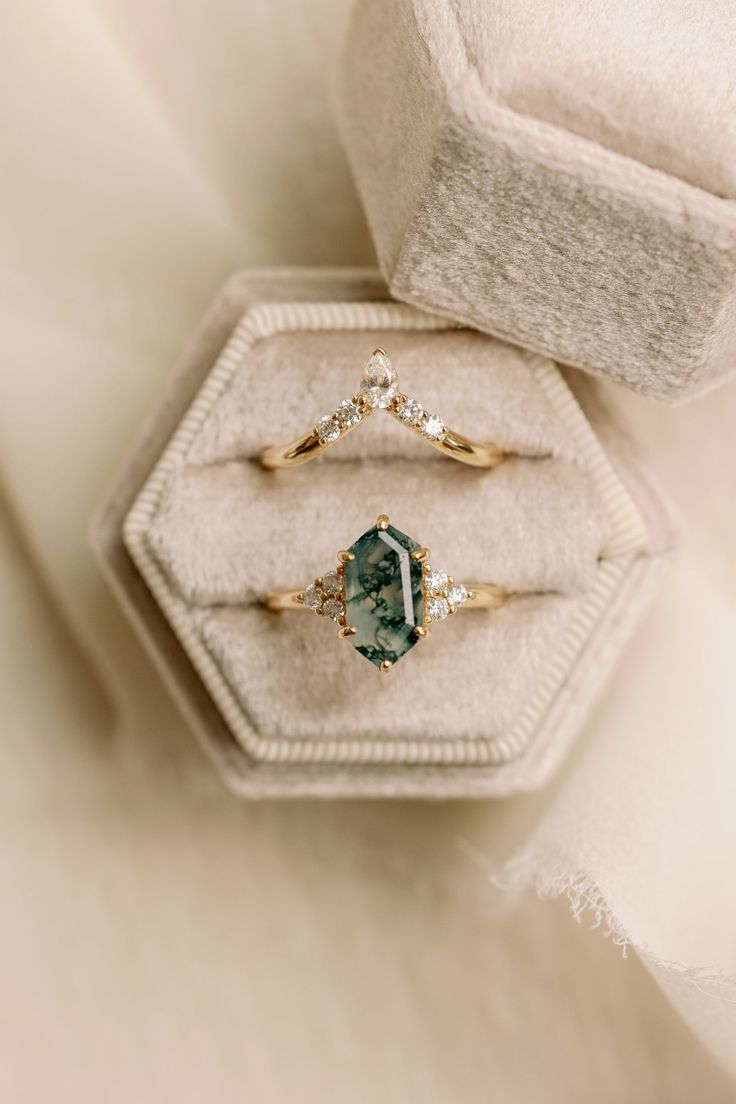 two engagement rings with an emerald and diamond set in a velvet ring box on top of a white furnishing