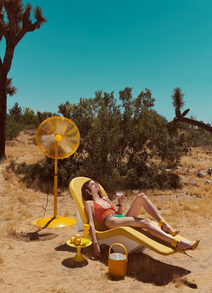 a woman lounging on a yellow lounge chair in the desert with a fan next to her