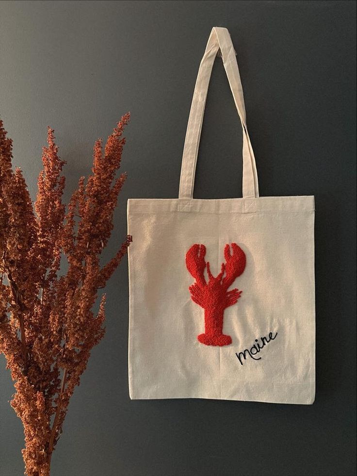 a white tote bag with a red lobster embroidered on it next to a plant