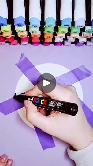 a person holding a marker in their hand near some markers on a purple table with different colored inks