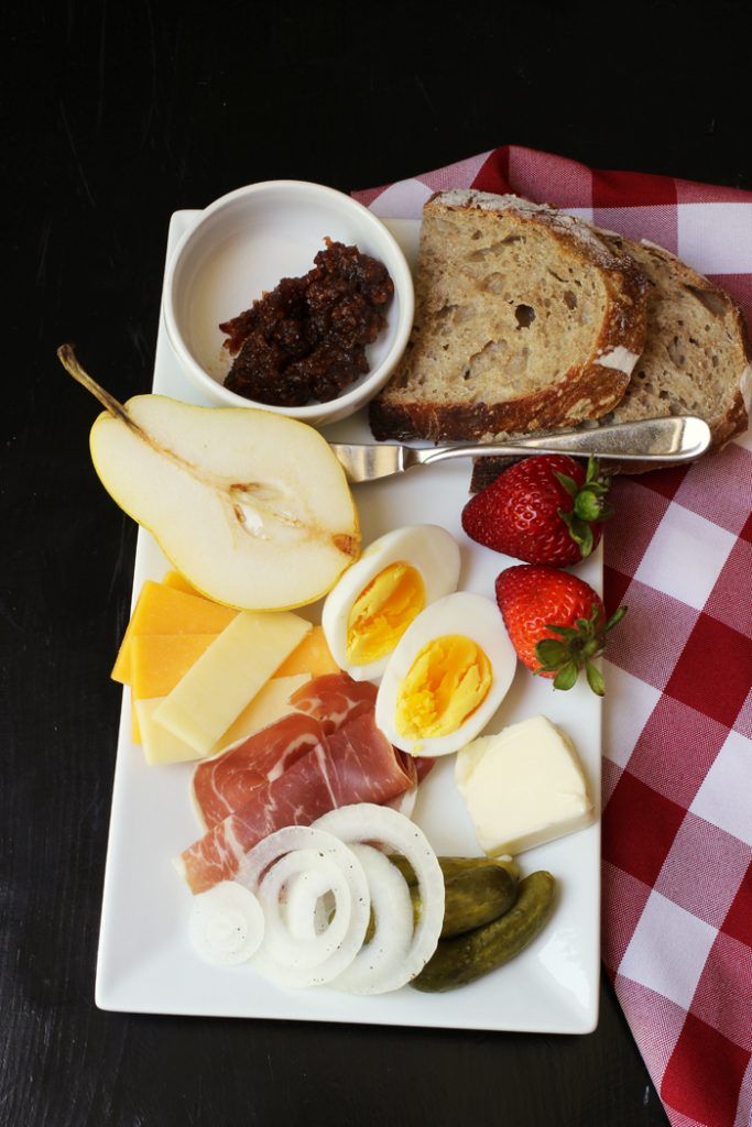 a white plate topped with meat, cheese and fruit next to a sandwich on a red and white checkered table cloth