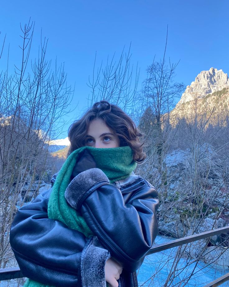 a woman wearing a green scarf and black jacket standing next to a river with mountains in the background