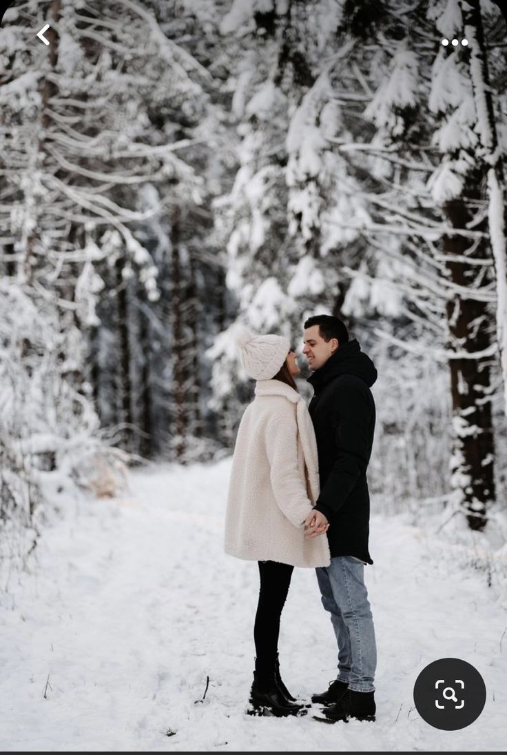 a man and woman standing in the snow near some trees with their noses to each other