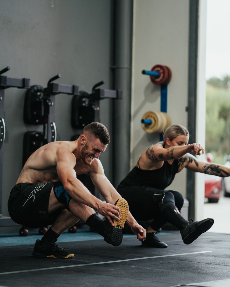 two men doing squats in a gym with one holding his leg up and the other sitting down