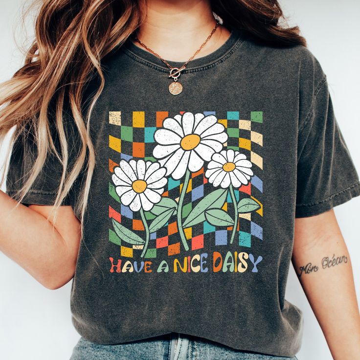 "Step into a cheerful vibe with our \"Have A Nice Daisy\" T-Shirt, a unique blend of retro aesthetics and comfort that is sure to bring a smile to your face. This shirt encapsulates a sense of nostalgia and positivity, making it a standout addition to your wardrobe. The shirt features the playful text, \"Have A Nice Daisy,\" designed in vibrant, retro colors, adding a pop of vibrancy and fun to any outfit. Sitting above the text is a beautifully illustrated daisy in retro style, its simple charm Daisy T Shirt Design, Retro Green Printed T-shirt, Casual Multicolor Vintage Print T-shirt, Retro Cotton Tops With Graphic Print, Black Casual Tops With Retro Print, Multicolor Retro Print Graphic Tee, Cotton Graphic Tee With Retro Print, Trendy Multicolor Funny Print Top, Retro Graphic Crew Neck Tee