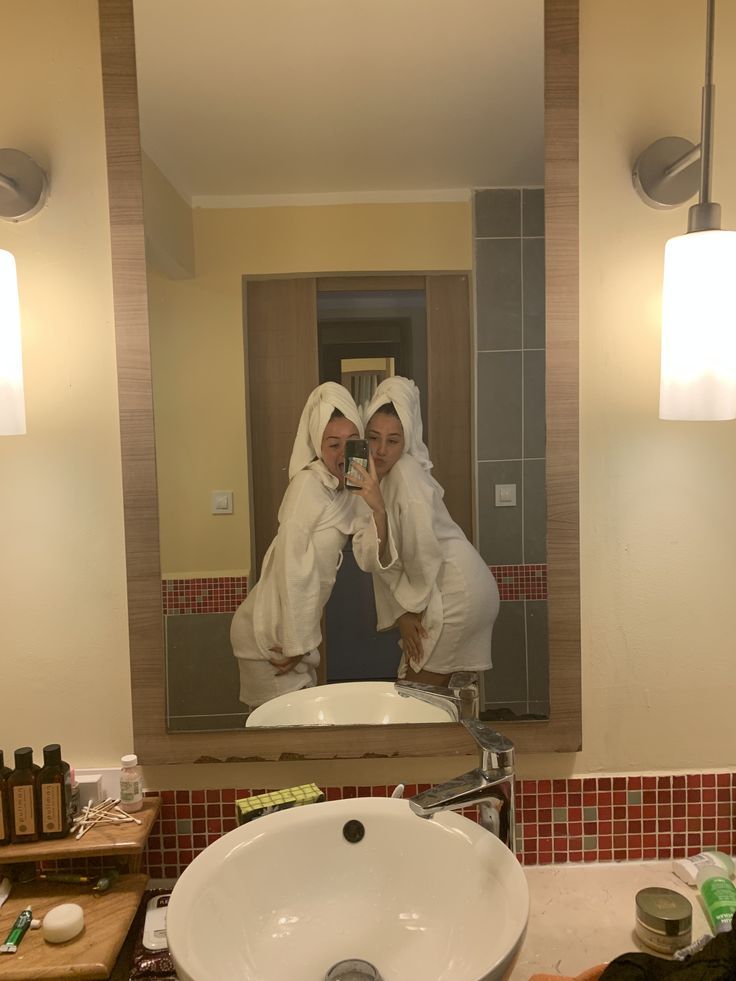 two women in white robes taking a selfie in a bathroom mirror with lights on