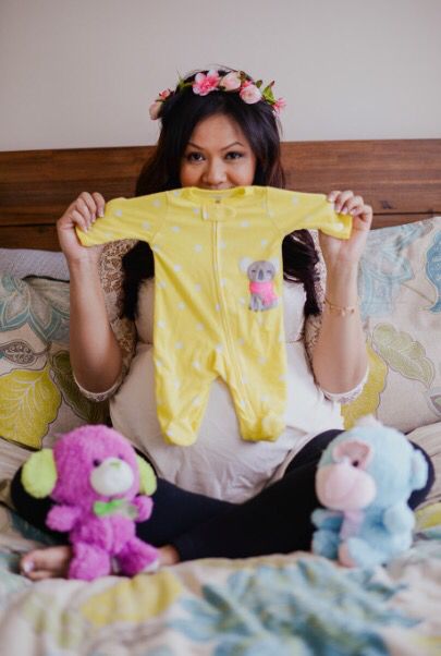 a woman sitting on a bed holding up a yellow baby onesuit and stuffed animals