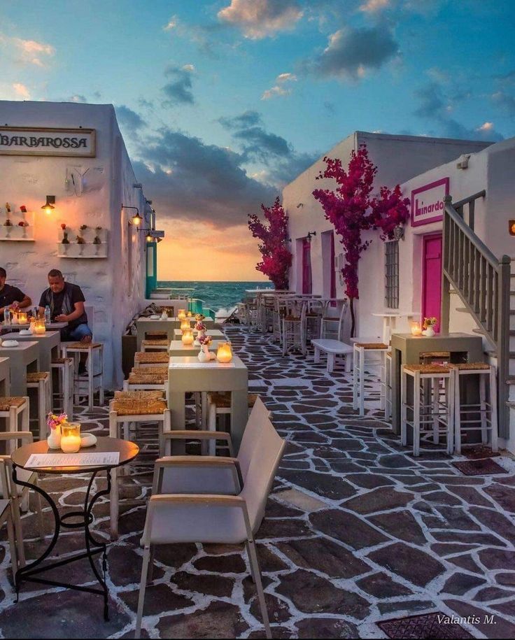 people are sitting at tables on the side of an alleyway with white buildings and red flowers