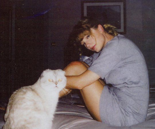 a woman sitting on a bed petting a white cat with her legs spread out