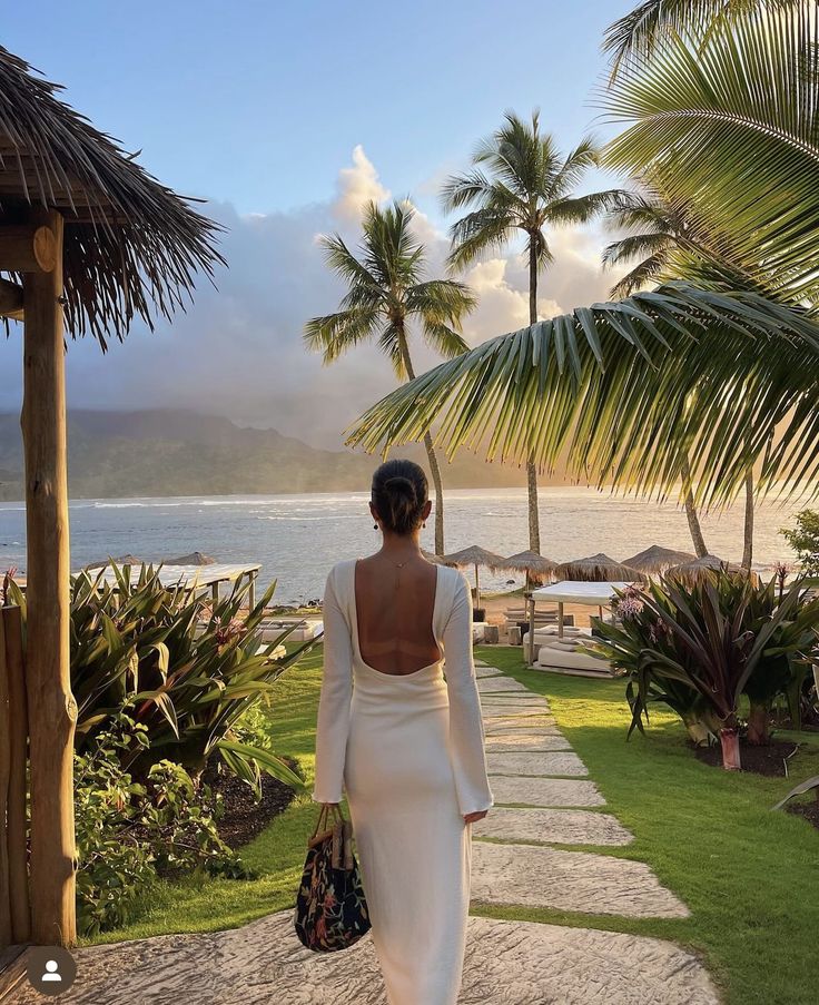 a woman in a white dress is walking towards the ocean with palm trees on either side