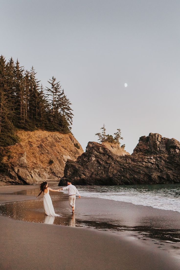 a man and woman walking on the beach with trees in the backgrouds