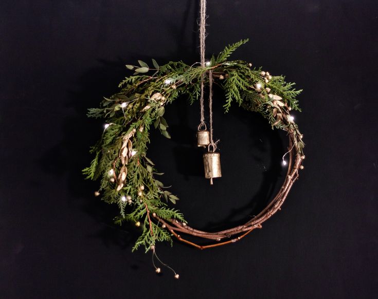 a christmas wreath with lights hanging from it's sides on a black wall next to two bells