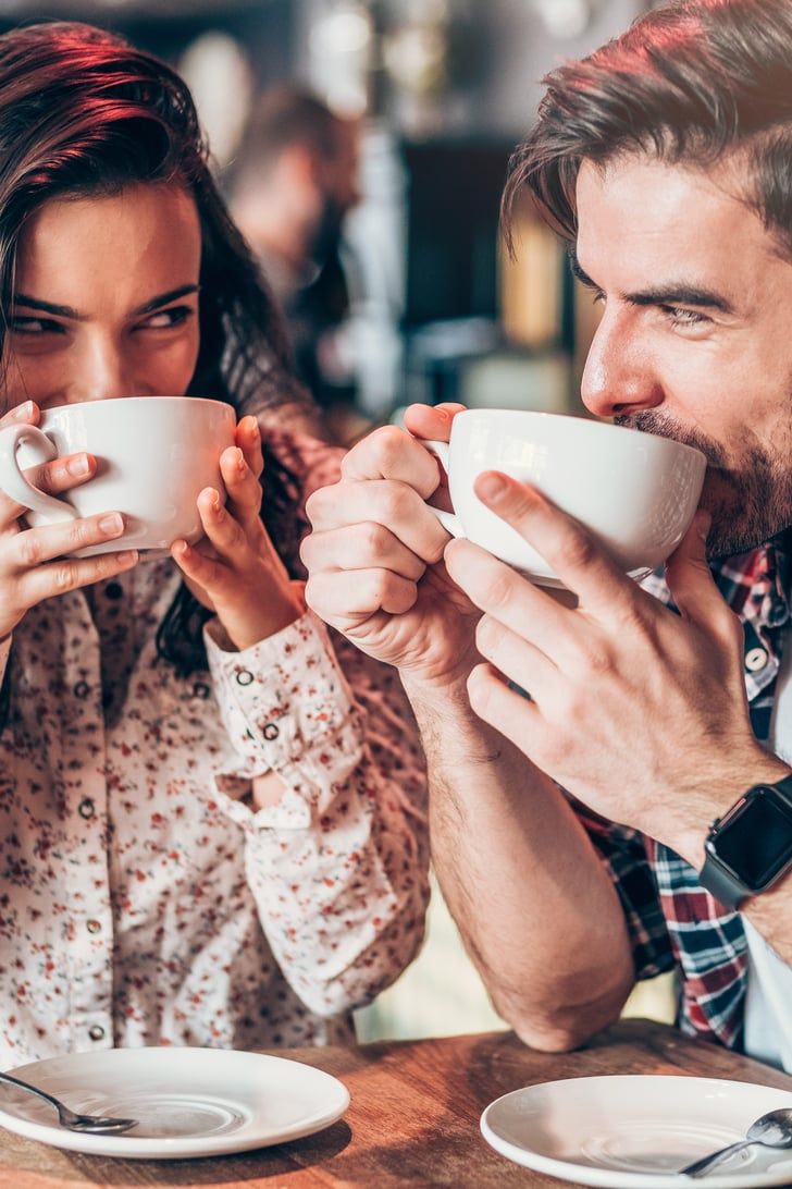 a man and woman sitting at a table with cups in front of their mouths, both holding coffee mugs