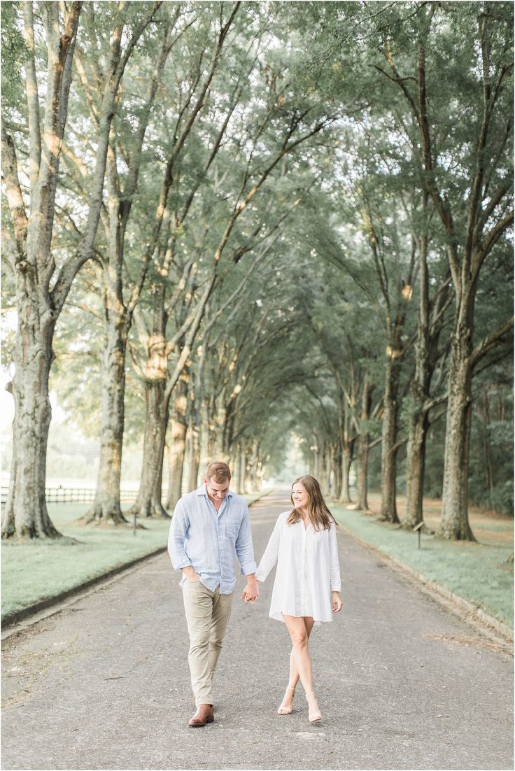 an engaged couple holding hands and walking down the road in front of some tall trees