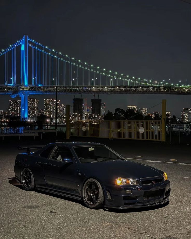 a black car parked in front of a bridge at night with the city lights behind it