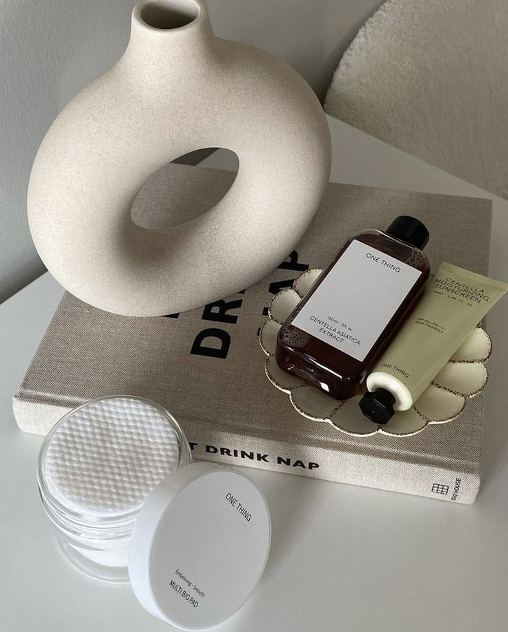 A bedside table with a funky vase resting on a stack of books. There’s a glass dishes filled with perfume bottles and cotton pads in a plastic container off to the side. Insta Content Ideas Lifestyle, Skincare Lifestyle Aesthetic, Skin Care Room Decor, Beige Skincare Aesthetic, Beauty Product Photoshoot Ideas, Beauty Content Creator Aesthetic, Ugc Content Aesthetic, Self Care Photoshoot Ideas, Ugc Content Creator Aesthetic