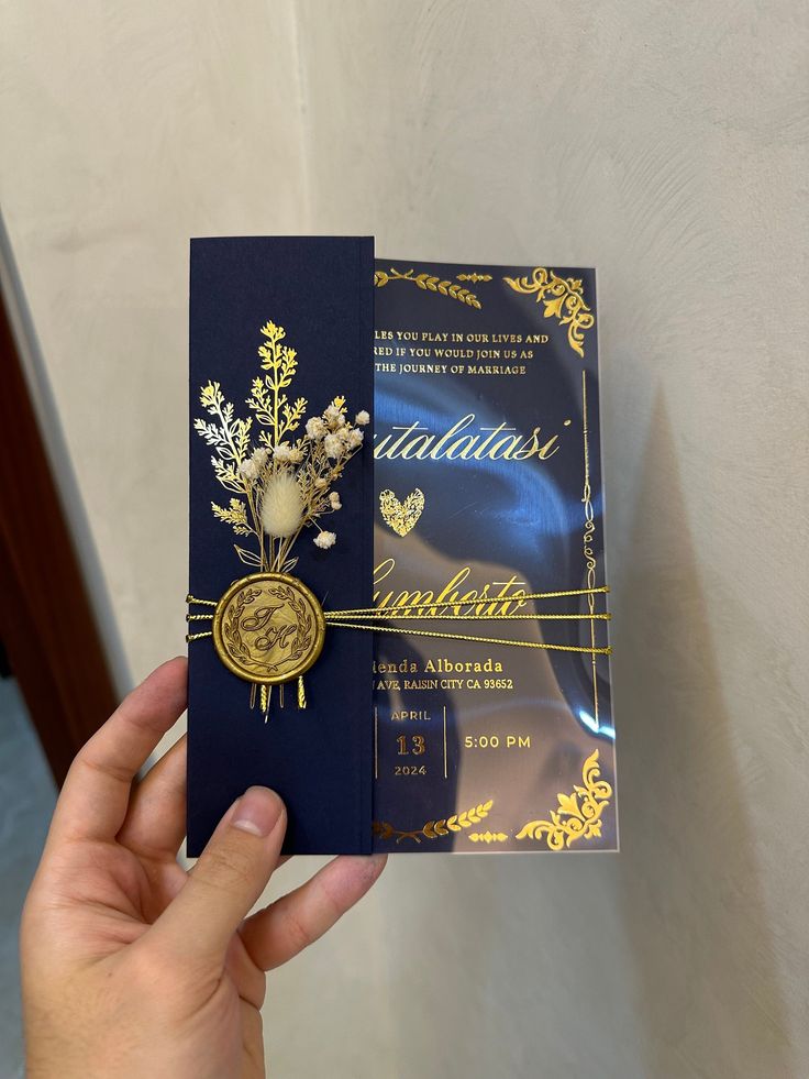 a person holding up a blue and gold wedding card with flowers on the front side