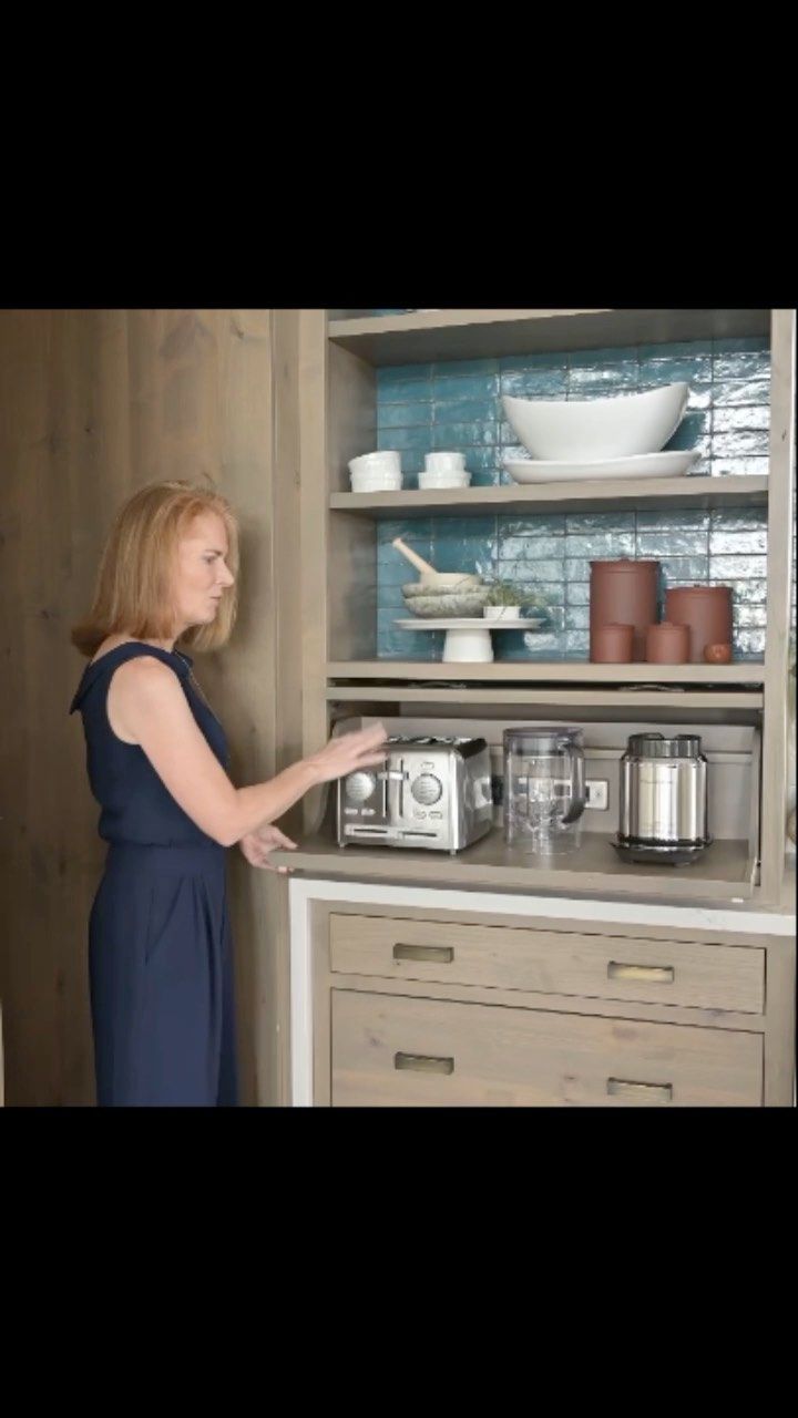 a woman standing in front of a cabinet with many items on it and she is pointing at the microwave