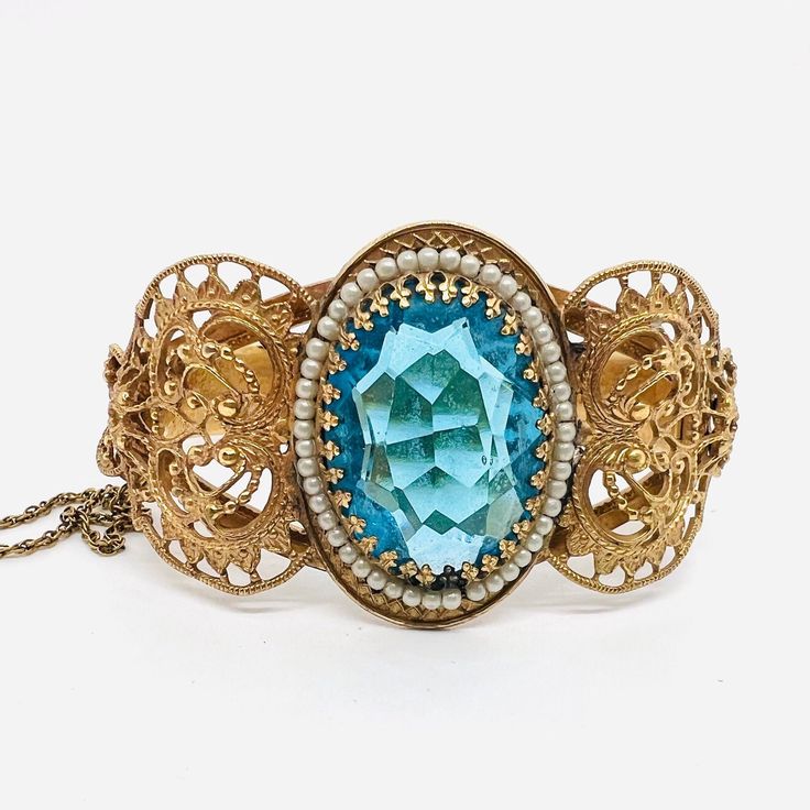 Gorgeous Vintage/Antique Wide Ornate Victorian Revival Blue Topaz Glass Bangle Bracelet. Gold tone/plated ornate filigree metal set with large topaz glass stone surrounded by faux seed pearls. In excellent vintage condition with minimal age appropriate wear. Measures 1 3/8 inches wide in from and 1/2 inch wide in back. Opening is 2 1/4 inches across. Has secure push in clasp and it's original safety chain Bohemian Jeweled Bracelets For Formal Occasions, Bohemian Jeweled Bracelet For Formal Occasions, Blue Intricate Design Jewelry Bracelet, Ornate Gold Bracelets With Jewels, Ornate Jeweled Bracelets For Formal Occasions, Ornate Metal Bracelets With Jewels, Ornate Jeweled Gold Bracelet, Ornate Gold Jeweled Bracelets, Ornate Gold Jeweled Bracelet