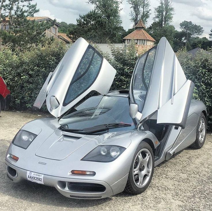 a silver sports car with its doors open in front of some bushes and trees on a gravel road