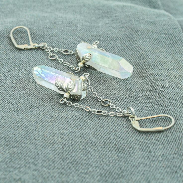 These beautiful angel aura quartz earrings are a dazzling addition to your jewelry collection. The rainbow glimmer on the crystal shimmers as the light catches it at different angles. Each crystal has two hand molded flower charms that wrap around the crystal and the silver finish give them an elegant yet bold feel. These are real gemstone earrings that dangle and have some weight to them. These earrings are not for those who are sensitive to heavily weighted earrings, but if you like to flaunt Silver Crystal Earrings With Gemstones, Mystical Silver Jewelry With Matching Earrings, Silver Dangle Jewelry With Mineral Crystal, Ethereal Silver Dangle Jewelry, Nickel-free Silver Jewelry With Mineral Crystal, Silver Dangle Mineral Crystal Jewelry, Mystical Silver Jewelry With Ear Wire, Mystical Silver Crystal Earrings For Gifts, Silver Mystical Jewelry With Ear Wire