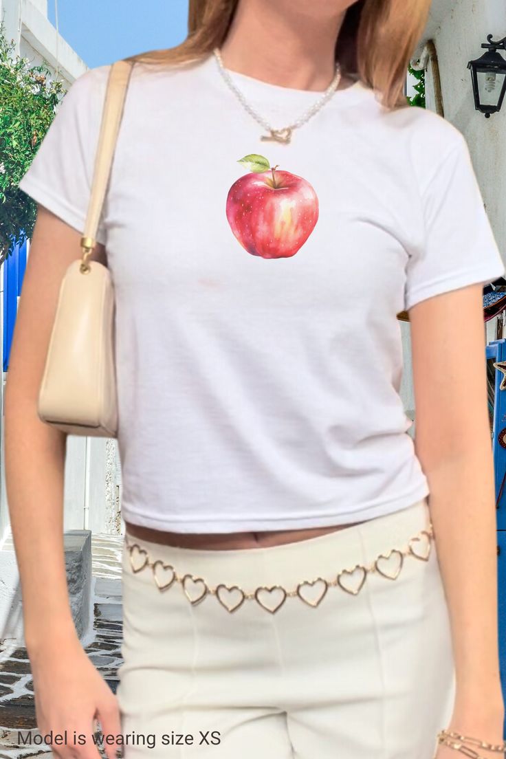 An apple a day keeps the doctor away 🍎 Get your fruity vitamins while wearing our trendy y2k baby tee shirt with a graphic red apple print. Style it with your low rise jeans or skirt and y2k sunglasses for the perfect vintage 2000s fit. Get yours now and wear it yourself or get a matching gift for your bestie in crime!  ------------------------------------------------------------------------------------------------------ Size guide Please take a moment to see the size chart, both in inches and Y2k Style Short Sleeve T-shirt With Fruit Print, Summer Y2k T-shirt With Fruit Print, Cute White Tops With Fruit Design, Cute White Fruit Design Tops, Y2k Fruit Print Short Sleeve Tops, Y2k Short Sleeve T-shirt With Fruit Print, Y2k Style Fruit Print Short Sleeve Tops, Y2k Short Sleeve Top With Fruit Print, Fitted Short Sleeve T-shirt With Fruit Print