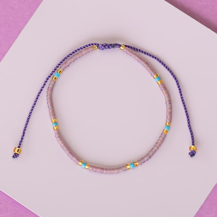 a purple beaded bracelet with gold beads on a white card next to a pink envelope