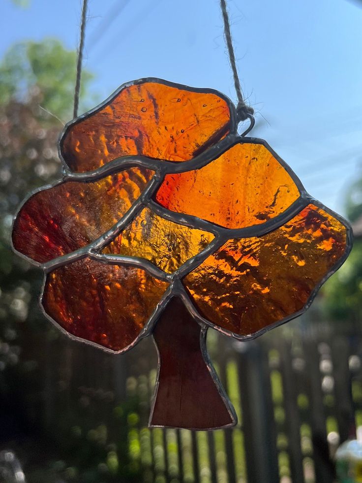 an orange stained glass flower hanging from a string in front of a fence and trees