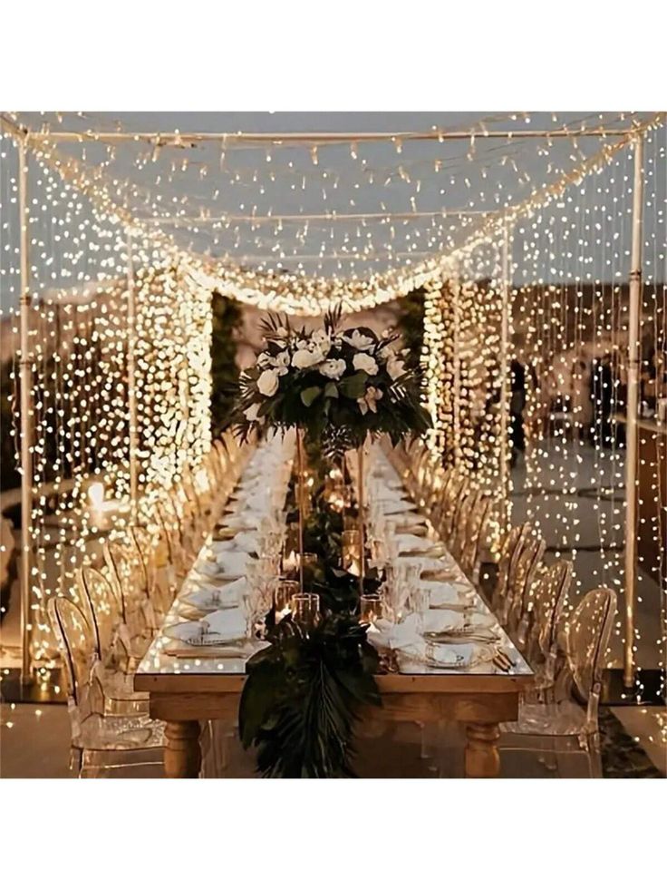 a long table covered in white lights and greenery with flowers on top is surrounded by clear chairs