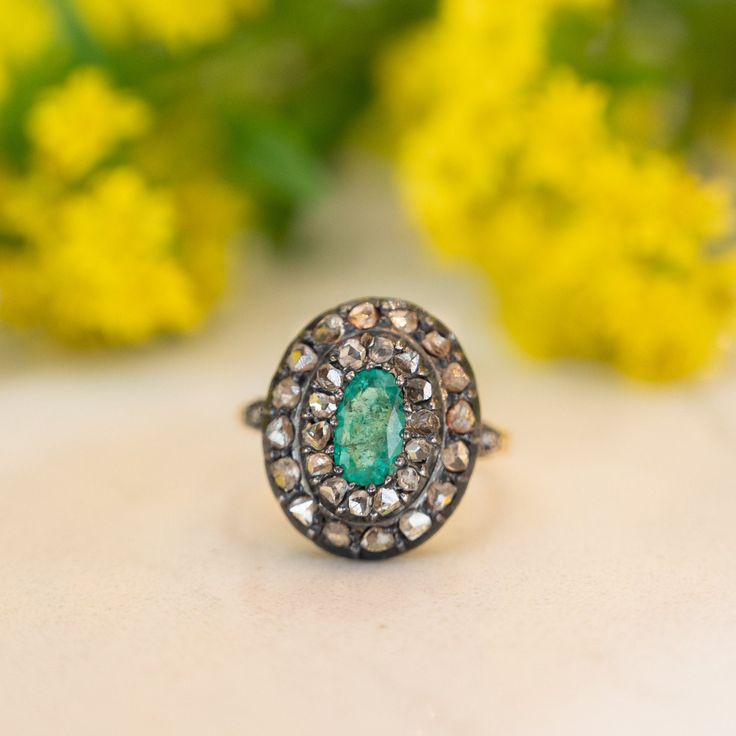 a ring with an oval shaped green stone surrounded by small white and clear stones, sits on a table next to yellow flowers