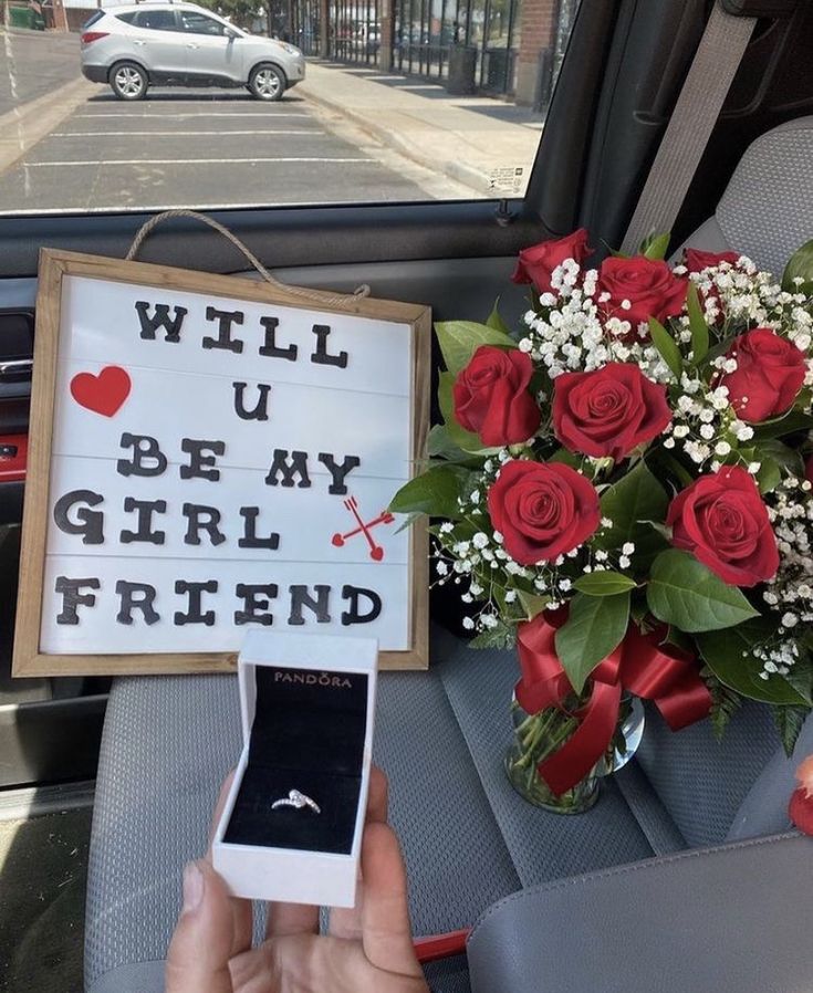 a person holding a cell phone next to a bouquet of roses and a sign that says will u be my girlfriend