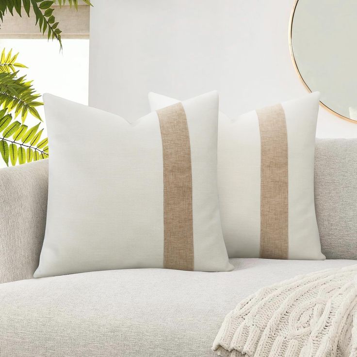 two white and beige pillows on a couch next to a potted plant in front of a mirror