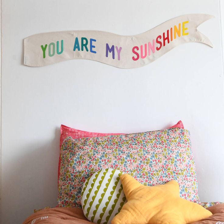 a bed topped with pillows next to a wall mounted sign that says you are my sunshine